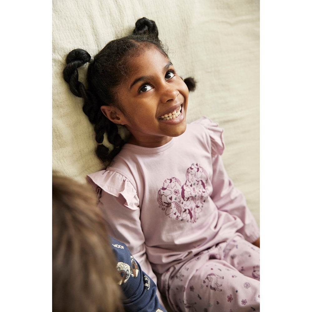 Mädchen trägt Pyjama von Minymo aus einer weichen Baumwollmischung bestehend aus einem langärmeligen T-Shirt und einer langen Hose, die einen All-Overprint mit Einhörnern hat. Das T-Shirt hat Rüschen an der Schulter und Stofffalten auf der Brust und ein Herz auf der Brust, das mit Einhörnern ausgefüllt ist.