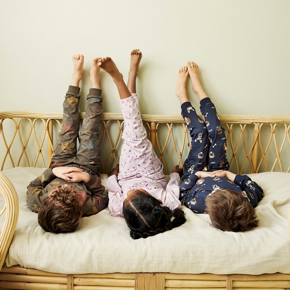 Drei Kinder tragen Pyjamas von Minymo. Einmal in braun mit Bagger, einmal in rosa mit Einhörnern und einmal in dunkelblau mit Hunden.